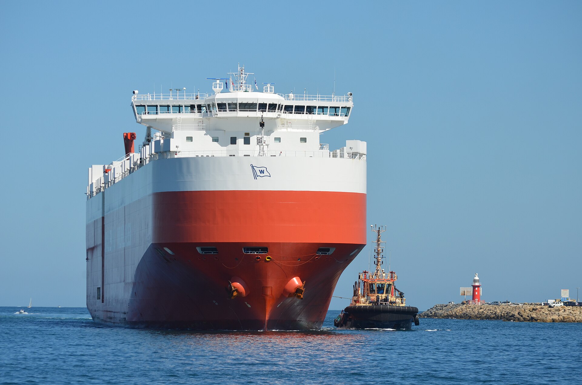 Een groot autoschip met een sleepboot ernaast bij de havenmond van Fremantle