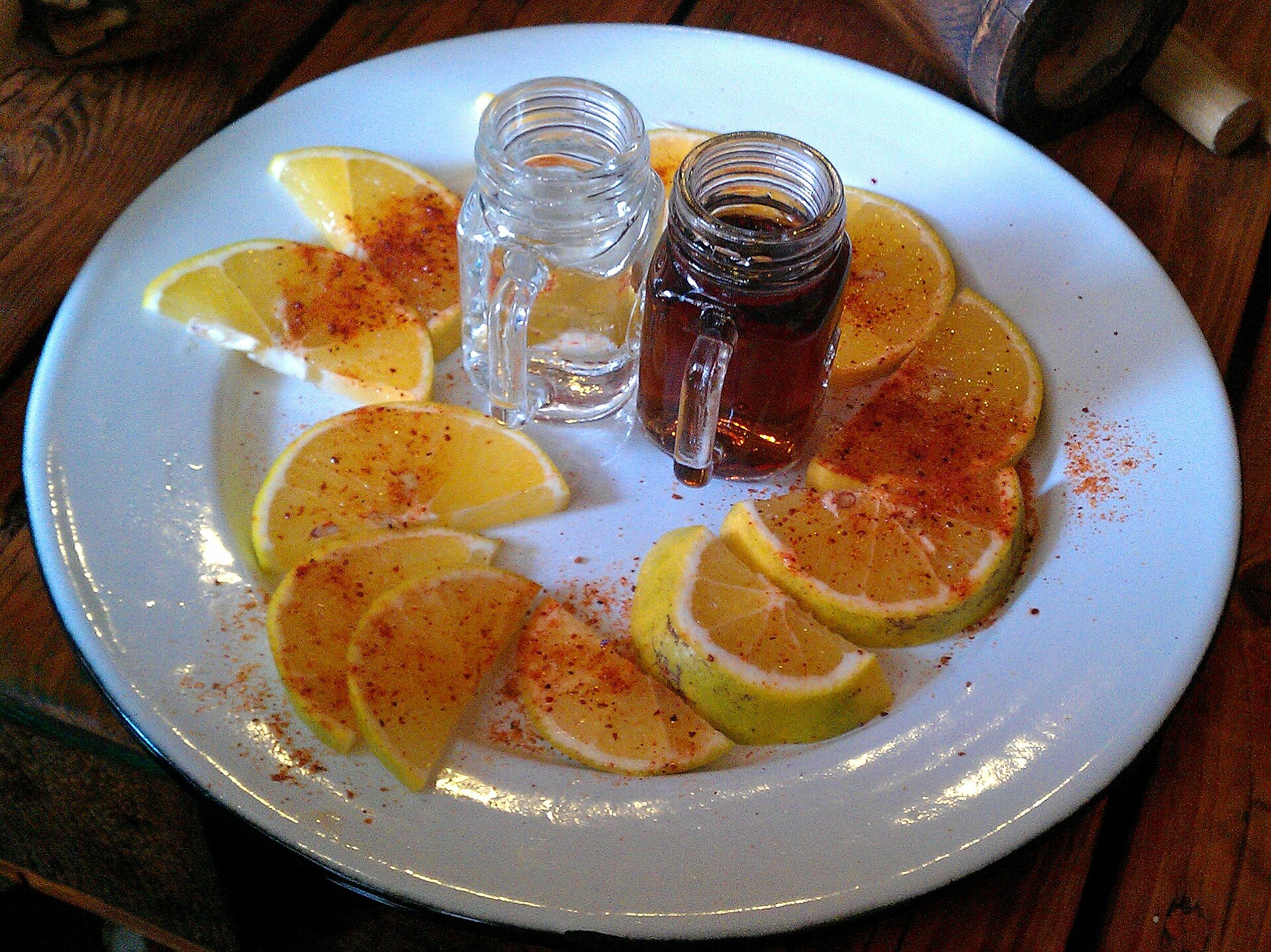 Een bord met schijfjes sinaasappel bestrooid met zout van gemalen larven, met twee glaasjes mezcal ernaast