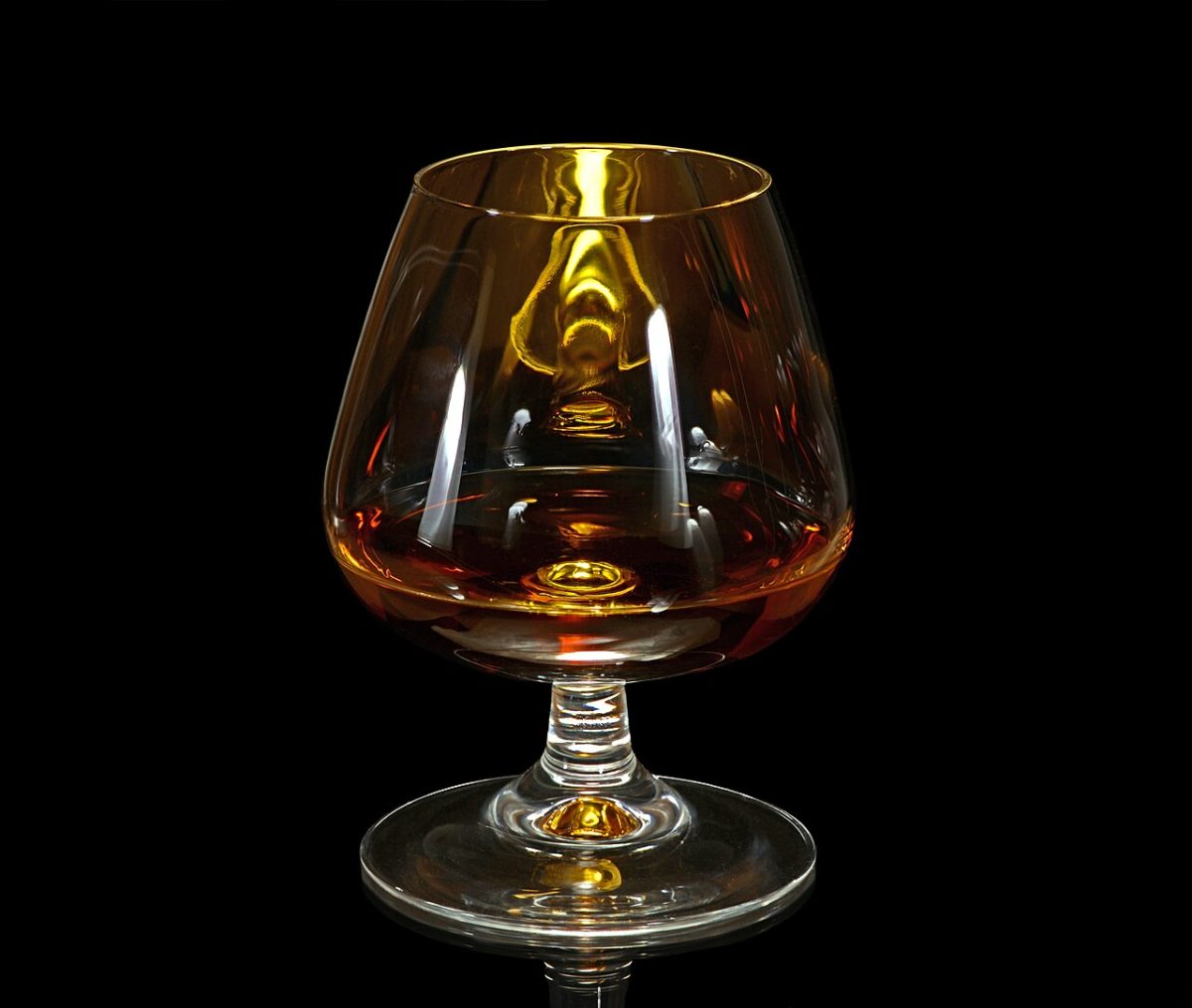 Ein bauchiges Cognacglas mit Stiel und bernsteinfarbenem Getränk vor schwarzem Hintergrund