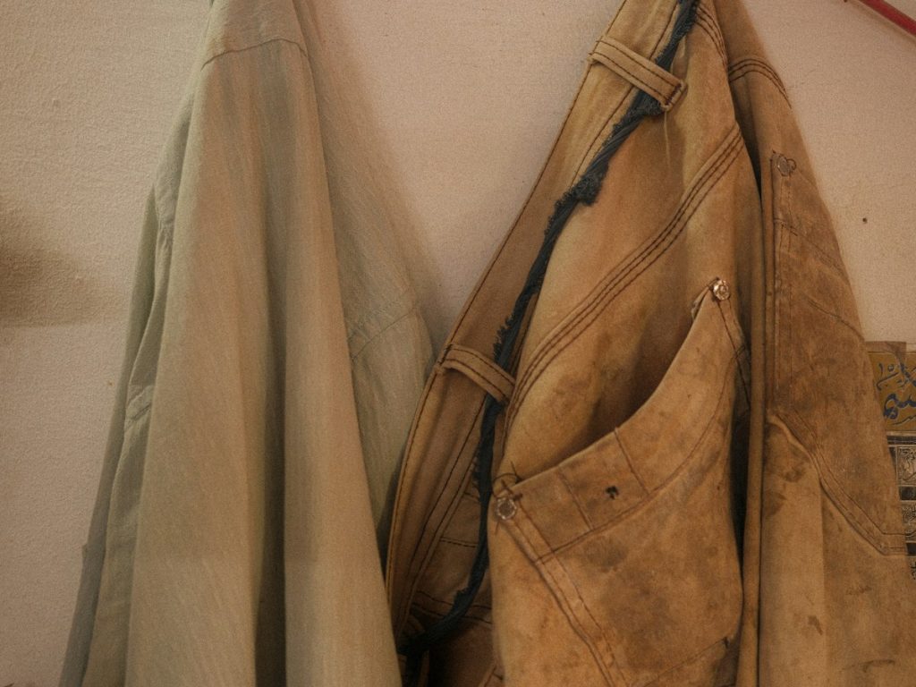 Worn workwear: a work jacket and work trousers on a hook against a light wall