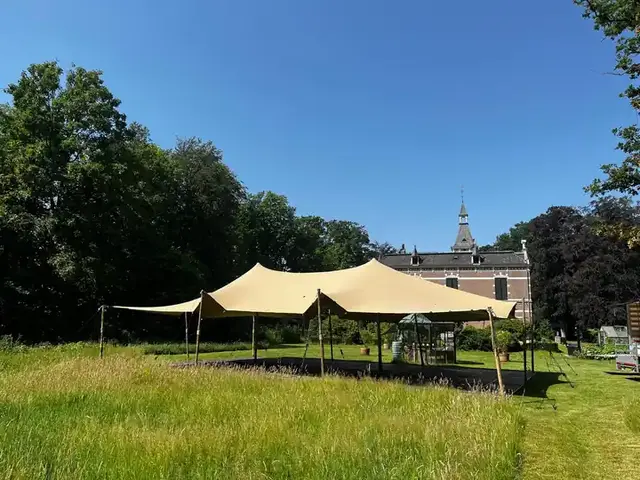 Zandkleurige stretchtent op houten palen in een parktuin bij een landhuis
