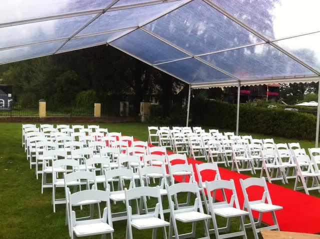 Tent met transparant dak boven rijen witte klapstoelen en een rode loper